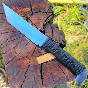 May include: A fixed-blade knife with a silver-colored tanto-style steel blade and a handle wrapped in dark gray cord. The blade features a small engraved star symbol near the hilt, and the handle ends in a rounded, polished metal pommel.