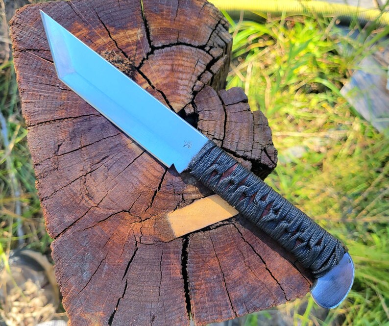 May include: A fixed-blade knife with a silver-colored tanto-style steel blade and a handle wrapped in dark gray cord. The blade features a small engraved star symbol near the hilt, and the handle ends in a rounded, polished metal pommel.