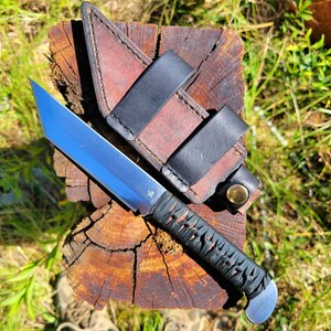 May include: A fixed-blade knife with a silver tanto-style steel blade and a black cord-wrapped handle. The knife is accompanied by a brown leather sheath featuring black leather straps and a brass-colored metal snap button closure.