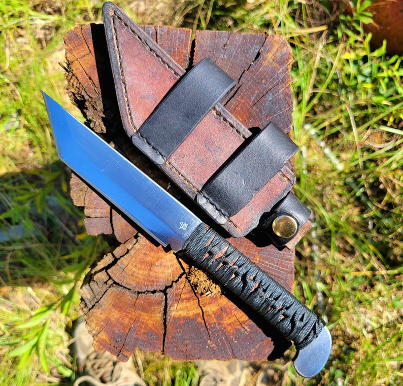May include: A fixed-blade knife with a silver tanto-style steel blade and a black cord-wrapped handle. The knife is accompanied by a brown leather sheath featuring black leather straps and a brass-colored metal snap button closure.