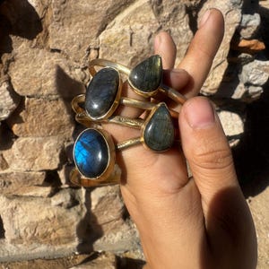 May include: A set of three gold-toned metal cuff bracelets, each featuring a single gemstone. Two bracelets have oval-shaped dark stones, and one bracelet has a teardrop-shaped stone. The metal bands display a subtle hammered or textured finish.