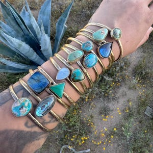 Può includere: Una collezione di braccialetti a polsino in metallo dorato, ognuno ornato da una gemma unica. Le gemme variano per forma e colore, tra cui turchesi, blu, verdi e pietre iridescenti. I braccialetti sono impilati su un polso.
