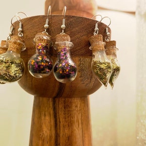 May include: Clear glass bottle earrings with cork stoppers. Some bottles contain gold glitter, others have purple and orange glitter, and some have green leaf-like pieces. The earrings hang from silver hooks and are displayed on a wooden stand.