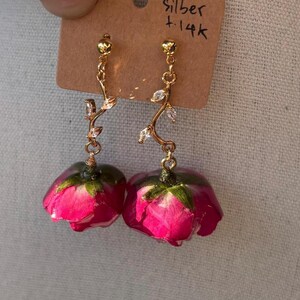 Puede incluir: Un par de pendientes colgantes dorados con un tallo en forma de enredadera con pequeños cristales transparentes, unidos a un colgante de capullo de rosa rosa. Presentados en una tarjeta marrón con el texto escrito a mano Silber +14k.