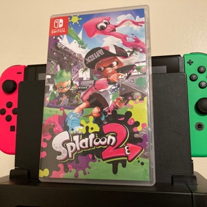May include: A Nintendo Switch console with a pink and green controller attached. The console is sitting on a black surface with a video game case on top. The game case is for Splatoon 2 and features a colorful design with a squid and two characters.