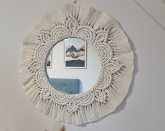 May include: A round, cream-colored macrame mirror with a detailed, fringed border. The mirror reflects a room with artwork and a blue headboard. The macrame design features intricate knotting patterns, adding a bohemian touch to the wall decor.