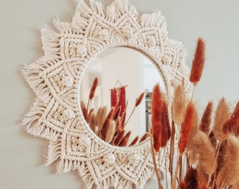 May include: A circular wall mirror framed by intricate off-white macrame cord featuring geometric patterns, knotted details, and a fringed outer edge. The macrame design creates a sunburst effect around the central glass mirror surface.