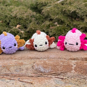 Peut inclure: Trois peluches d'axolotl au crochet, alignées. Les axolotls sont de couleurs violet, blanc et rose, avec des branchies et des accents contrastés. Ils ont de petits yeux noirs et sont posés sur une surface en pierre.