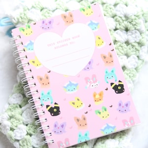 Puede incluir: Un libro de pegatinas de espiral rosa con un patrón repetitivo de criaturas de dibujos animados coloridas y una etiqueta blanca en forma de corazón con el texto impreso THIS STICKER BOOK BELONGS TO y dos líneas en blanco.