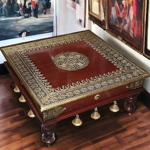 Könnte beinhalten: Ein quadratischer Holztisch mit einer roten Tischplatte und goldenen Verzierungen. Der Tisch hat vier Beine und ein dekoratives Design auf der Oberseite. Der Tisch ist mit Glocken verziert, die an der Unterseite hängen.