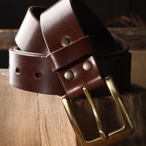 May include: A brown leather belt with a brass buckle. The belt is made of a smooth, dark brown leather and features a rectangular brass buckle. The belt is shown coiled on a wooden surface.