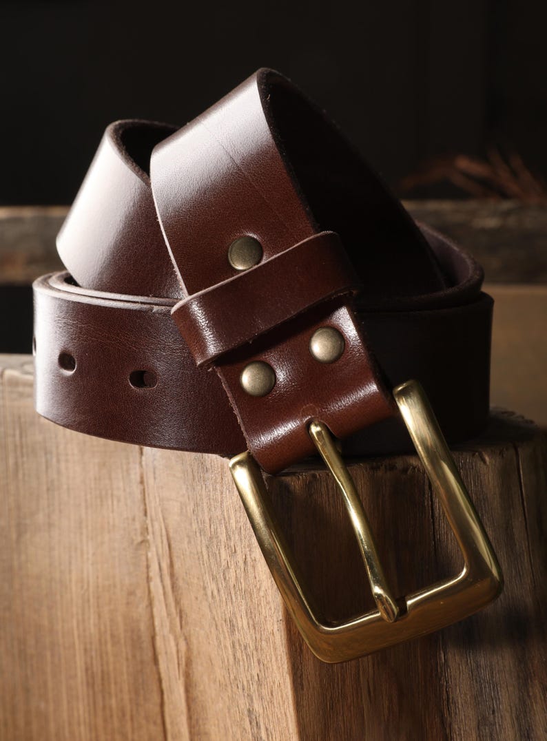 May include: A brown leather belt with a brass buckle. The belt is made of a smooth, dark brown leather and features a rectangular brass buckle. The belt is shown coiled on a wooden surface.