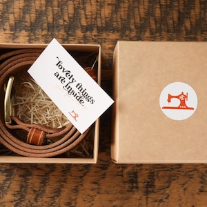 May include: A brown leather belt with a gold buckle is coiled inside a square box. A card with the text "lovely things are inside" rests on top. The box is next to a closed box with a sewing machine logo.