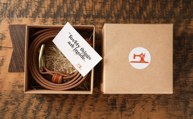 May include: A brown leather belt with a gold buckle is coiled inside a square box. A card with the text "lovely things are inside" rests on top. The box is next to a closed box with a sewing machine logo.