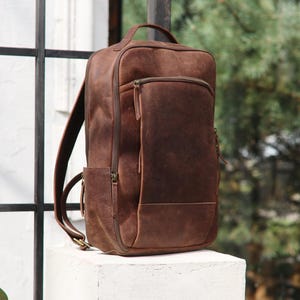 Peut inclure: Un sac à dos en cuir marron de forme rectangulaire avec une poignée supérieure. Le sac à dos a une poche avant zippée et une poche latérale. Les bretelles sont réglables. Le sac est posé sur une surface blanche.