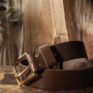 May include: A brown leather belt with a gold-colored buckle. The belt is rolled up, showcasing the rich color and texture of the leather. The buckle is rectangular and the belt has two brass rivets.