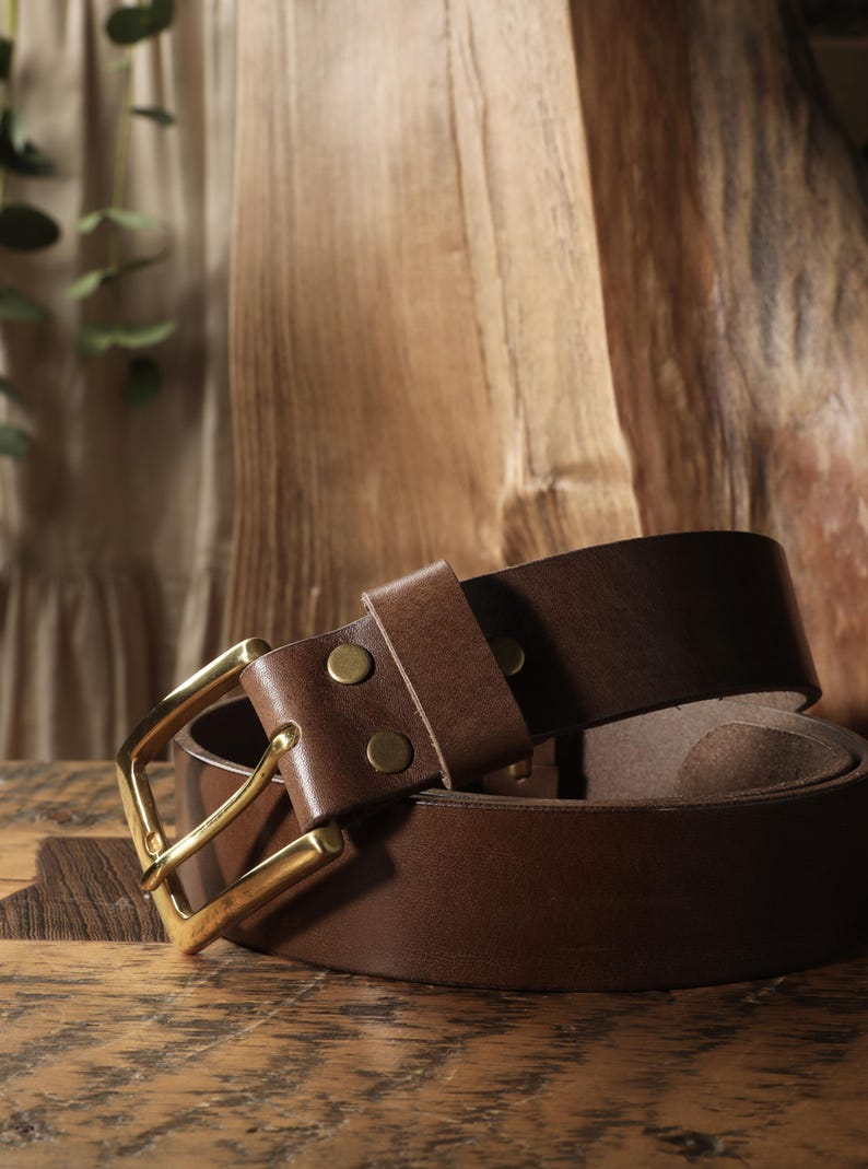 May include: A brown leather belt with a gold-colored buckle. The belt is rolled up, showcasing the rich color and texture of the leather. The buckle is rectangular and the belt has two brass rivets.