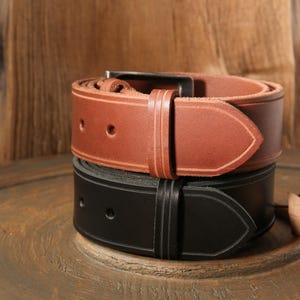 Peut inclure: Deux ceintures en cuir, une marron et une noire, superposées. La ceinture marron, avec une boucle argentée et des trous de réglage, est au-dessus de la ceinture noire. Les deux ceintures ont un design classique, largeur 3.8 cm.