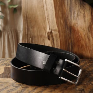 May include: A black leather belt with a silver-colored buckle. The belt is rolled up on a wooden surface, showcasing its smooth texture and design. The buckle is rectangular and the belt has a classic, minimalist style.