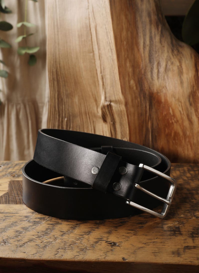 May include: A black leather belt with a silver-colored buckle. The belt is rolled up on a wooden surface, showcasing its smooth texture and design. The buckle is rectangular and the belt has a classic, minimalist style.