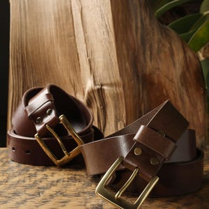 May include: Two brown leather belts with brass buckles are displayed on a wooden surface. The belts are rolled and unrolled, showcasing their design. The buckles have a rectangular shape.