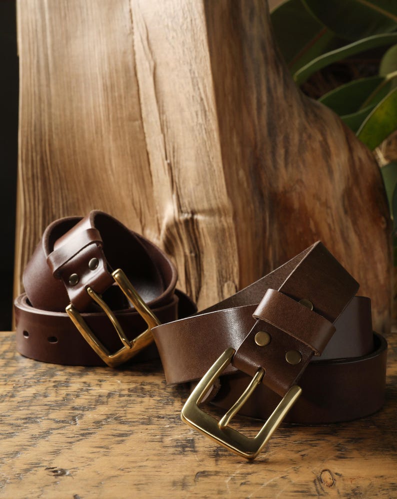 May include: Two brown leather belts with brass buckles are displayed on a wooden surface. The belts are rolled and unrolled, showcasing their design. The buckles have a rectangular shape.