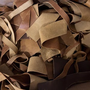 May include: A collection of leather offcuts in shades of brown and beige. The pieces are cut into strips and irregular shapes, with some curled and folded. The leather has a rough, worn texture, suggesting it's been used.