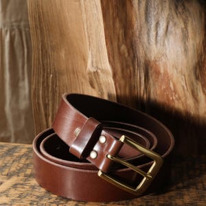 May include: A rolled brown leather belt with a gold-colored buckle. The belt is made of a smooth, dark brown leather and has a classic design. The buckle is rectangular and has a shiny finish.