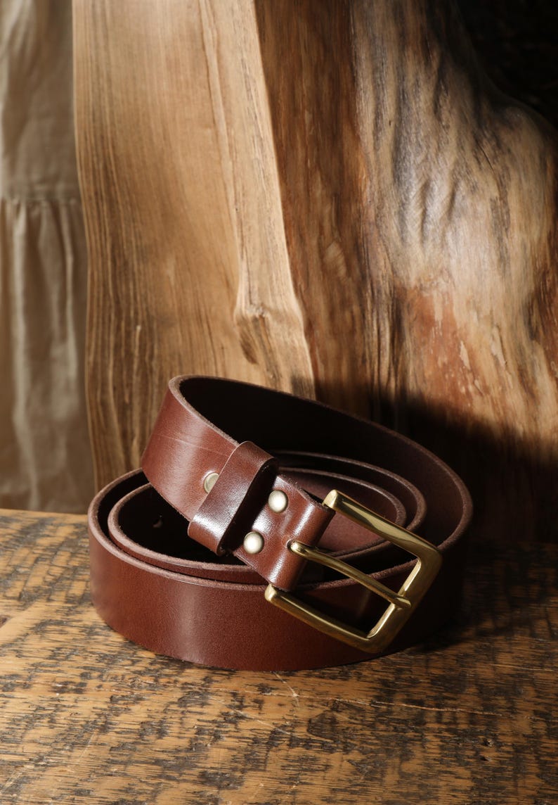 May include: A rolled brown leather belt with a gold-colored buckle. The belt is made of a smooth, dark brown leather and has a classic design. The buckle is rectangular and has a shiny finish.