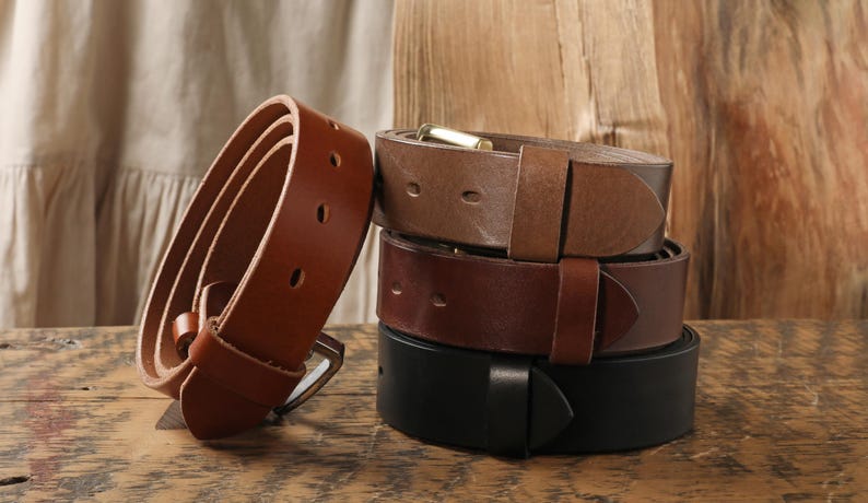 May include: A stack of four leather belts in shades of brown and black. One belt is coiled, showing the buckle and holes. The other three belts are stacked, showcasing their buckles and leather texture. The background is a wooden surface.