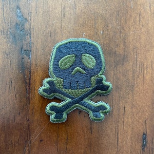Peut inclure: Un patch brodé vert et noir représentant un crâne et des os croisés. Le crâne a des yeux verts et un contour noir.