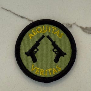 Peut inclure: Un écusson brodé vert et noir avec les mots "Aequitas Veritas" et deux pistolets croisés en fil jaune.
