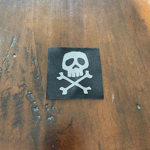 Könnte beinhalten: Ein schwarzer Stoff-Patch mit einem weißen Totenkopf- und Knochen-Design.