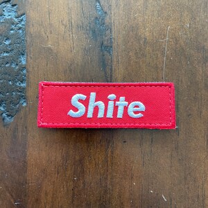 Könnte beinhalten: Roter Stoff-Patch mit weiß gesticktem Text, der "Shite" lautet.