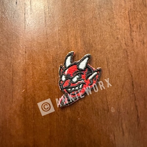Puede incluir: Un parche de diablo rojo y negro con cuernos blancos y el texto "kinkieworx". El parche está sobre una superficie de madera. El parche es un diseño bordado.