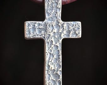 May include: A silver cross pendant with a textured surface. The pendant is hanging from a brown leather cord.
