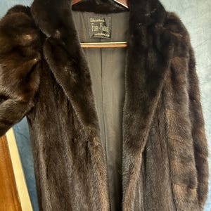 Puede incluir: Un abrigo de piel marrón oscuro con cuello ancho y mangas largas. El abrigo está colgado en una percha de madera. La etiqueta dice "Winston's Fur-Faire".