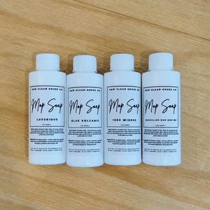 May include: Four white plastic bottles of Raw Clean House Co. Mop Soap, each with a white screw cap and a label featuring black text. The labels specify the scents Luxurious, Blue Volcanic, 1000 Wishes, and Brazilian Bum God Me, with 2 oz sample text.