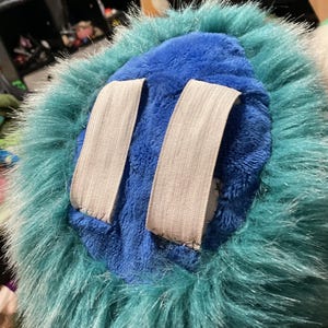 Puede incluir: Un juguete de peluche azul con un ribete de piel turquesa. El juguete tiene dos correas el&aacute;sticas blancas en la parte posterior.