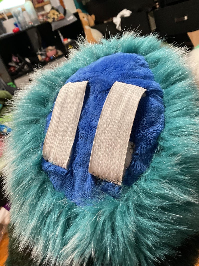 Puede incluir: Un juguete de peluche azul con un ribete de piel turquesa. El juguete tiene dos correas el&aacute;sticas blancas en la parte posterior.