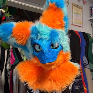 May include: A full-head animal mask featuring bright orange and light blue faux fur. The face is molded in a textured blue material with large, mesh-covered eye openings and a prominent snout. The ears are tall, pointed, and covered in fur.