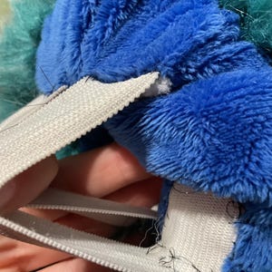 Puede incluir: Primer plano de un juguete de peluche azul y verde con correas el&aacute;sticas blancas cosidas alrededor del borde. Las correas est&aacute;n cosidas con hilo negro.