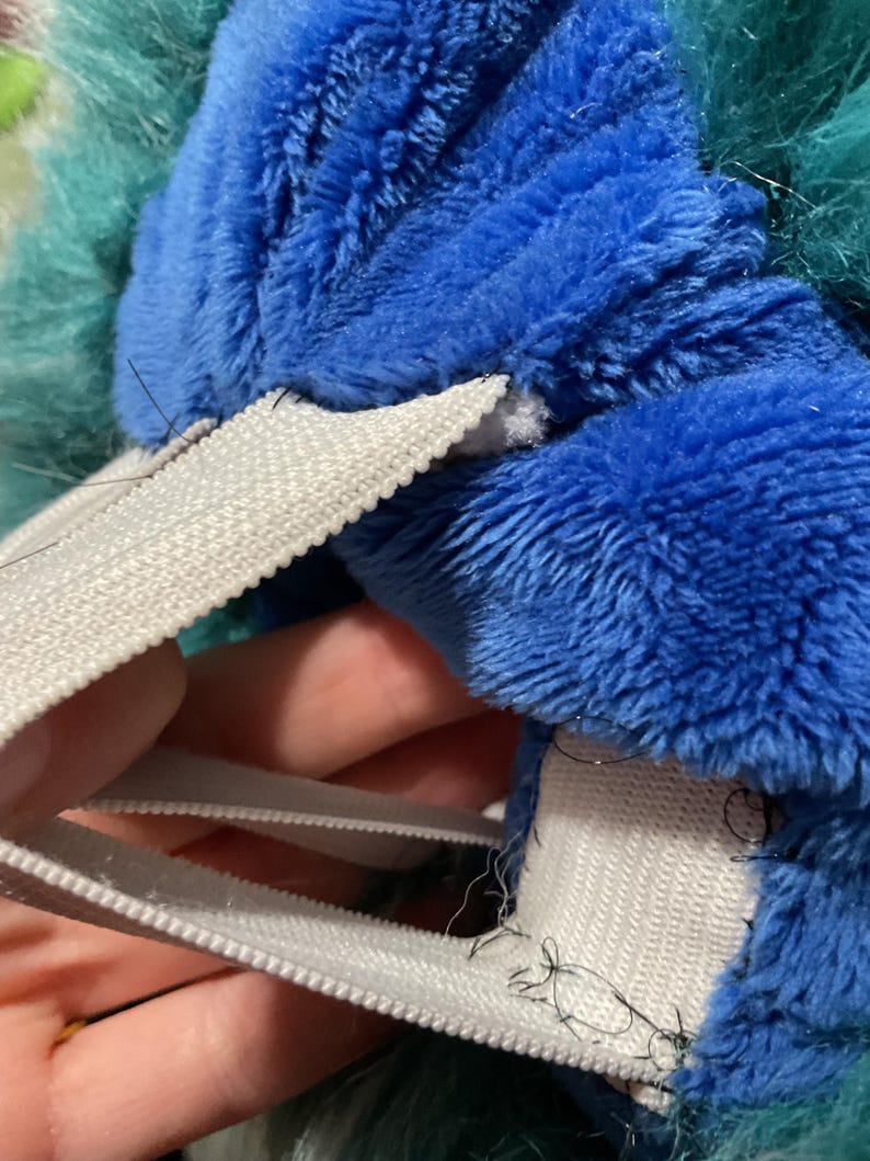 Puede incluir: Primer plano de un juguete de peluche azul y verde con correas el&aacute;sticas blancas cosidas alrededor del borde. Las correas est&aacute;n cosidas con hilo negro.