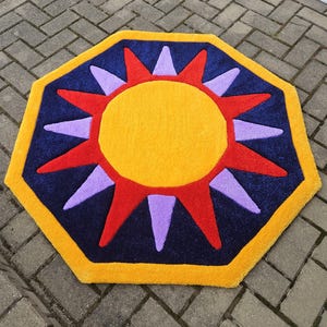Può includere: Un tappeto a forma di sole giallo, rosso e viola su sfondo blu. Il tappeto è ottagonale e ha un bordo giallo.
