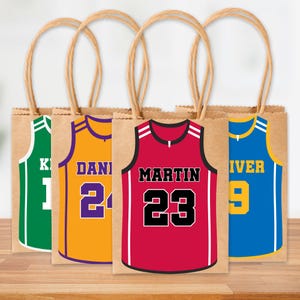 Könnte beinhalten: Vier braune Papiertüten mit Basketball-Trikot-Designs. Jede Tasche hat eine andere Trikotfarbe: grün, gold, rot und blau. Die Trikots zeigen Namen und Nummern, mit braunen Seilgriffen.