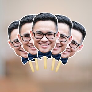 May include: A group of five face cutouts, each featuring a smiling person wearing glasses and a navy blue bow tie. The cutouts are attached to wooden sticks, ideal for photo booth props.