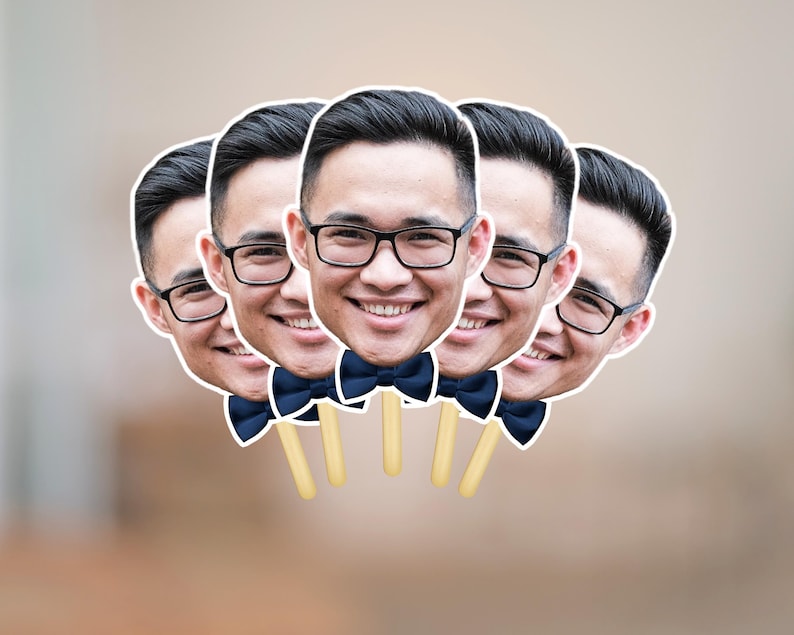 May include: A group of five face cutouts, each featuring a smiling person wearing glasses and a navy blue bow tie. The cutouts are attached to wooden sticks, ideal for photo booth props.