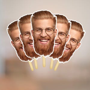 May include: A group of five face cutouts on sticks. Each cutout features a smiling man with a red beard, glasses, and light brown hair. The cutouts are white-bordered and set against a blurred background.