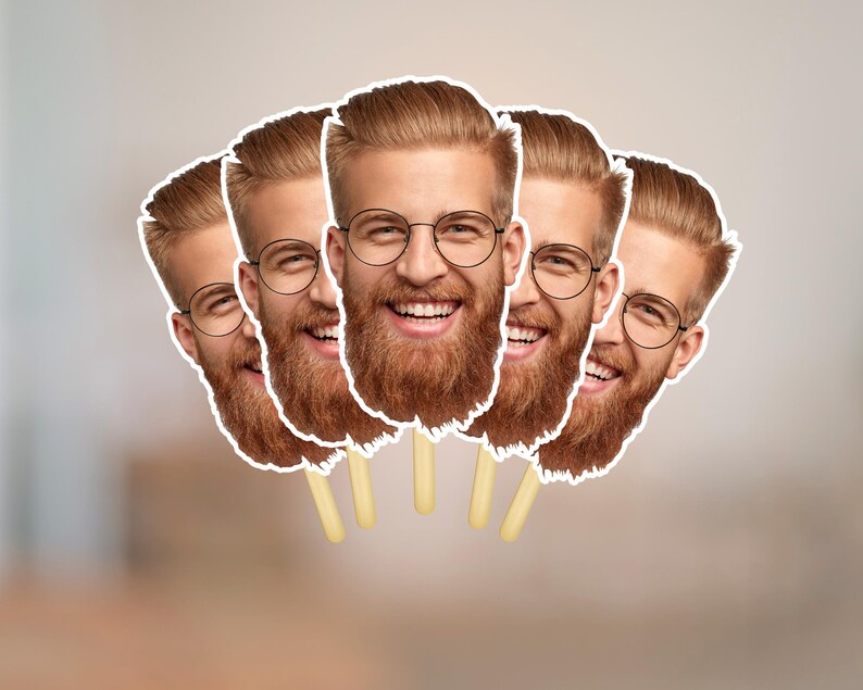 May include: A group of five face cutouts on sticks. Each cutout features a smiling man with a red beard, glasses, and light brown hair. The cutouts are white-bordered and set against a blurred background.