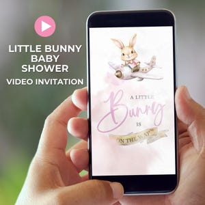 Op de afbeelding: Een smartphone toont een video-uitnodiging voor een babyshower. Het ontwerp toont een cartoonkonijn dat een roze vliegtuig bestuurt. De tekst luidt "Little Bunny Baby Shower Video Invitation" en "A Little Bunny Is On The Way."